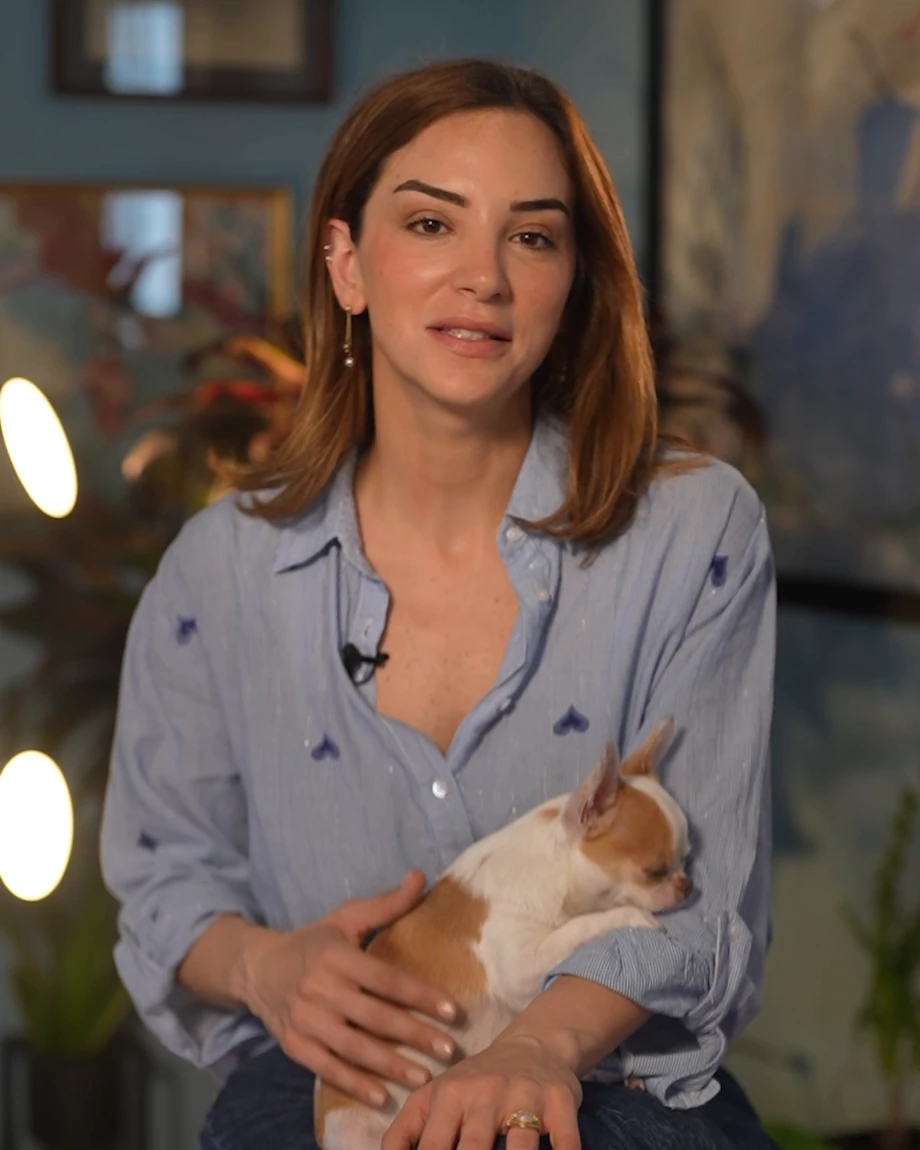 The artist at home in her atelier, holding her dog, with her artwork behind her