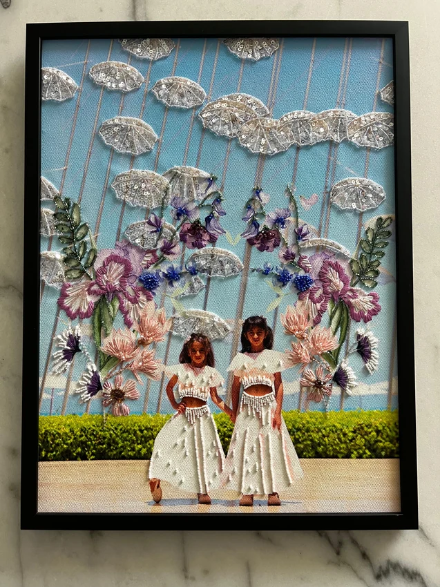 Two children beneath embroidered flowers and silver sequin clouds in a black frame