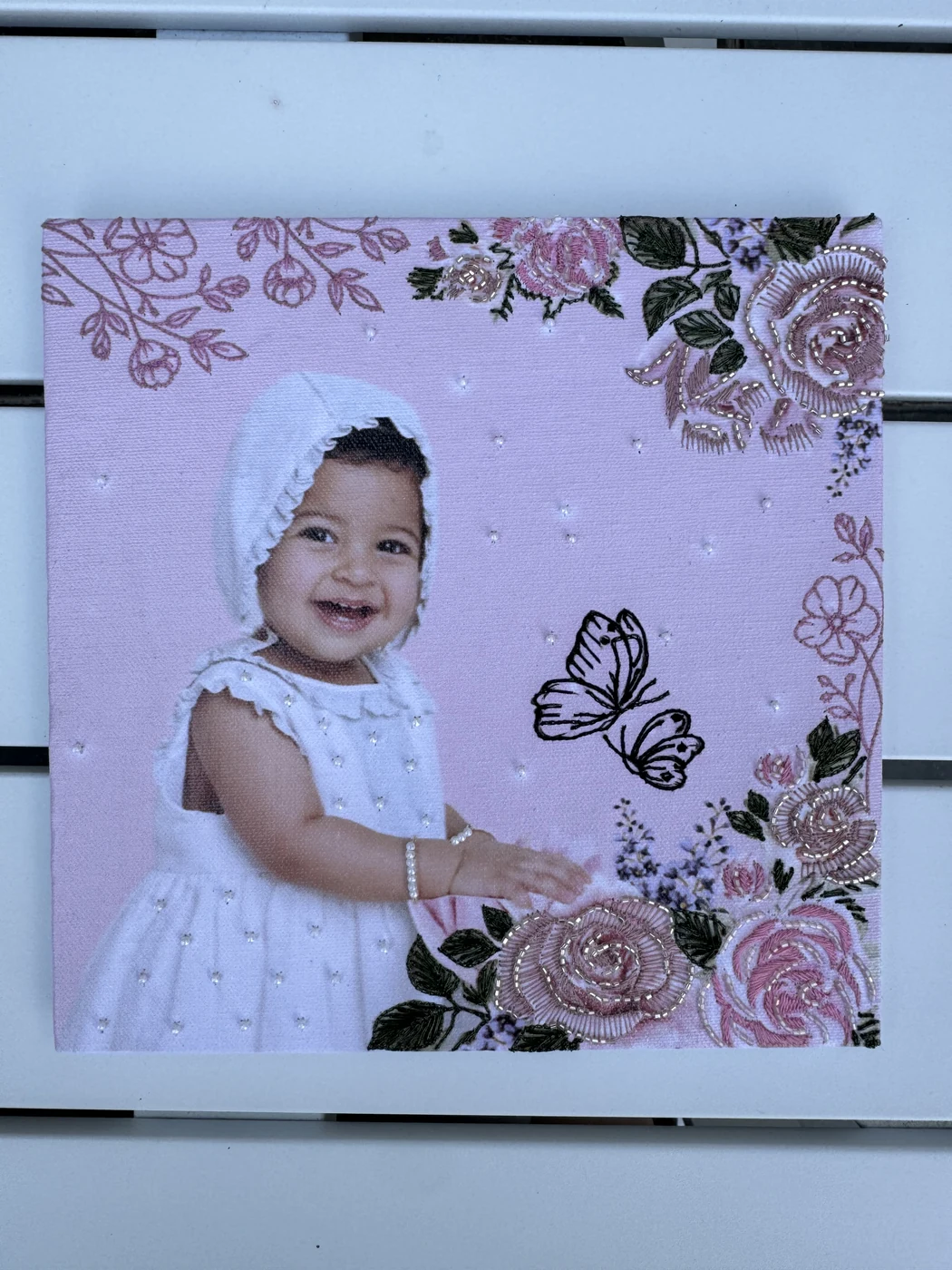 Smiling baby in a white dress surrounded by pink beaded roses, embroidered butterflies and pearls